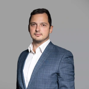Portrait photo of a man with short dark hair, wearing a blue suit and light shirt. He is looking at the camera with a serious expression, against a neutral grey background.