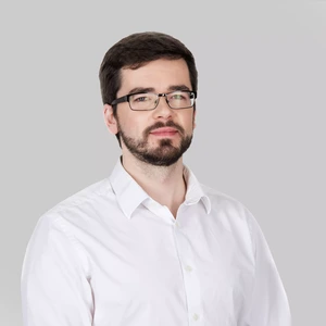 Serious-looking man with short dark hair and a beard, wearing a white shirt and glasses, grey background.