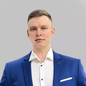 Young man with short light hair, wearing a bright blue suit and white shirt, looking directly at the camera, grey background.