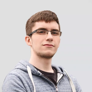 Portrait photo of a young man with short brown hair, wearing glasses, a grayish-blue hoodie, and a dark T-shirt underneath. He is looking at the camera with arms crossed against a neutral gray background.