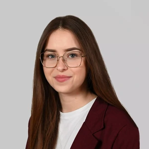 Portrait photo of a young woman with long, straight dark brown hair. She is wearing glasses, a white top, and a dark burgundy blazer. The background is light grey.
