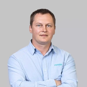 Portrait photo of a man with short dark hair, wearing a light blue shirt with the “Festo” logo. He is standing with arms crossed and looking at the camera with a serious, focused expression. The background is neutral light grey.