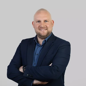 Portrait photo of a man with a shaved head, wearing a dark blue blazer and blue shirt. He is standing with arms crossed and smiling while looking at the camera. The background is neutral light grey.