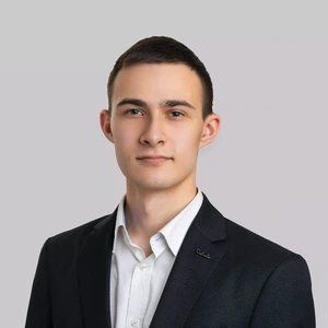 Portrait photo of a young man with short dark hair, wearing a dark blazer and a white shirt. He looks into the camera with a serious, neutral expression. The background is light gray and neutral.