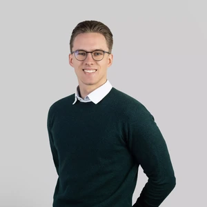 Portrait photo of a young man with short light hair, wearing a dark green sweater and white shirt, with glasses. He is looking at the camera with a wide smile, against a neutral grey background.