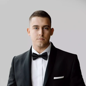 A young man with short hair, wearing a tuxedo with a bow tie and white dress shirt front, looks directly at the camera with a serious expression against a light background.