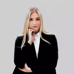 Portrait photo of a young woman with long blonde hair, wearing a black blazer and a white shirt, looking directly at the camera against a light background, resting her hand gently on her chin.