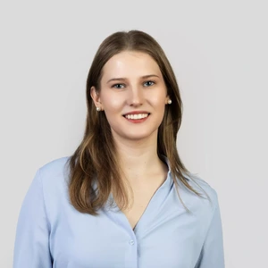 Portrait photo of a young woman with straight light brown hair, wearing a light blue shirt and pearl earrings. She is smiling while looking at the camera, with a light neutral background.