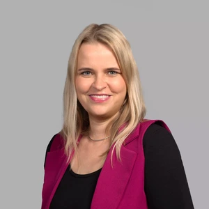 Portrait photo of a blonde woman with a gentle smile, wearing a black top and a bright pink vest. The background is neutral grey.
