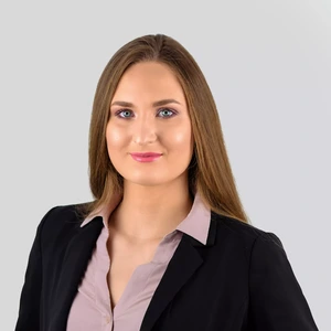 Portrait photo of a young woman with long, light brown hair. She is wearing a light pink blouse and a black blazer. The background is light grey.