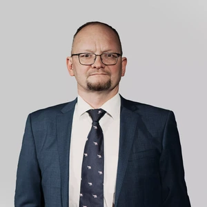A middle-aged man with glasses and a goatee is wearing a dark blue suit, white shirt, and a dark tie with small animal motifs. He looks directly at the camera with a serious expression against a light background.