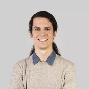 Portrait photo of a young man with long dark hair tied back in a ponytail. He is wearing a light sweater with a visible denim shirt collar. The man is smiling while looking at the camera, against a neutral light grey background.