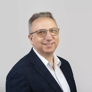 Portrait photo of a middle-aged man wearing glasses, a dark blue checkered blazer, and a white shirt, smiling while looking at the camera against a neutral gray background.
