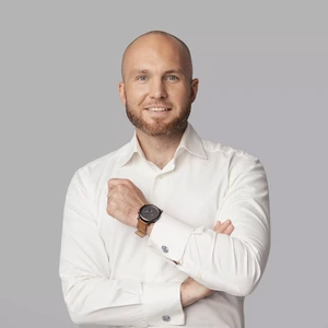 Portrait photo of a man with a neatly trimmed beard and a shaved head, wearing a white shirt. He is smiling at the camera, with a watch visible on his right wrist. The background is neutral grey.