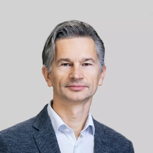 Portrait photo of a middle-aged man with greying hair, wearing a grey blazer and a light shirt, looking directly at the camera against a neutral grey background.