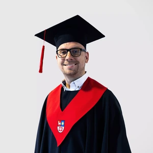 Portrait photo of a man with short hair and glasses, wearing academic regalia with a bright red collar accent and a dark academic cap with a red tassel. He is smiling while looking at the camera. The background is neutral light grey.