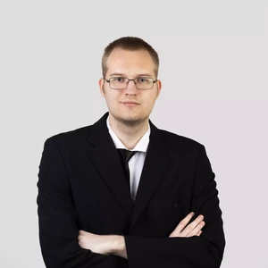 A short-haired man wearing glasses and a black suit, standing with arms crossed against a light background.