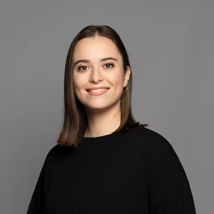 Portrait photo of a woman with short dark hair, wearing a black blouse. She is smiling and looking at the camera against a gray background.