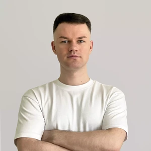 Portrait photo of a young man with short dark hair, wearing a plain white shirt. He stands with arms crossed and looks into the camera with a serious expression. The background is light gray and neutral.