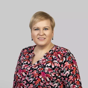 Portrait photo of a woman with short blonde hair, wearing a colorful patterned blouse featuring pink and red floral motifs. She is looking at the camera with a pleasant smile. The background is neutral light grey.