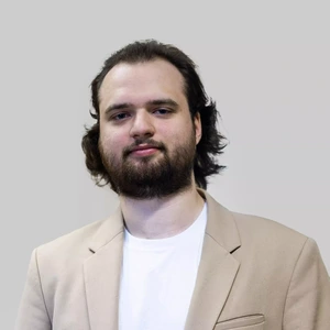 Portrait photo of a young man with a full beard and medium-length dark hair. He is wearing a beige blazer and a white t-shirt, looking directly at the camera with a subtle smile. The background is neutral grey.