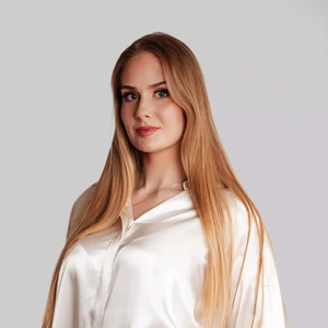 A young woman with long blonde hair, wearing a white silk blouse, looks directly at the camera with a calm and confident expression against a light background.