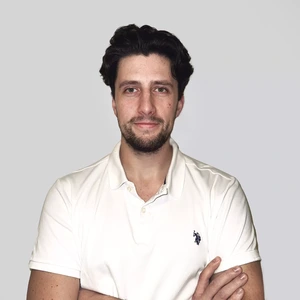 Portrait photo of a young man with dark wavy hair and a beard, wearing a white polo shirt, looking at the camera with arms crossed against a light background.