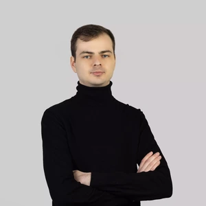 Portrait photo of a young man with short hair, wearing a black turtleneck. He is standing with arms crossed and looking directly at the camera with a neutral facial expression. The background is light grey and neutral.