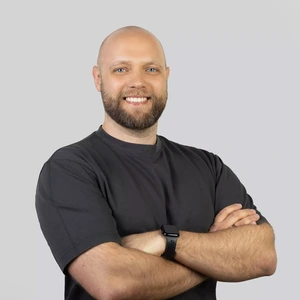 A bald man with a short beard, wearing a dark T-shirt, standing with arms crossed and smiling.