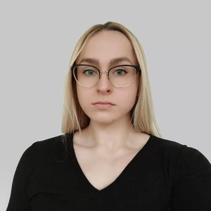 Portrait photo of a young woman with light hair, wearing glasses and a black top, looking directly at the camera against a light background.
