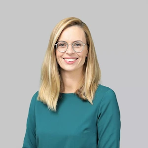 A woman with blonde hair and round glasses, wearing a green blouse, is smiling and looking directly at the camera against a light background.