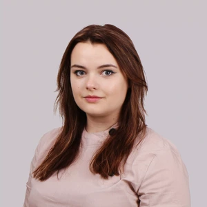 Portrait photo of a young woman with brown hair, wearing a light pink top, looking at the camera against a neutral grayish background. A small microphone clipped to her top suggests the photo might have been taken during an interview or public presentation.