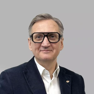 Portrait photo of an older man wearing thick-rimmed glasses, a white shirt, and a dark blazer. He is smiling and looking directly at the camera. The background is grey.