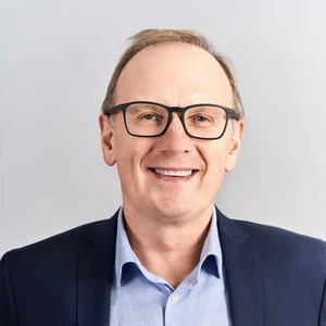 Portrait photo of an older man with glasses and light hair, wearing a light blue shirt and a dark blue blazer. He is smiling and looking directly at the camera. The background is neutral, light grey.