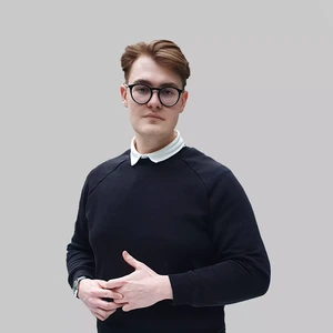 Portrait photo of a young man with short blond hair, wearing a black sweater and a white shirt with a collar. He is looking at the camera with a serious expression, against a neutral grey background.