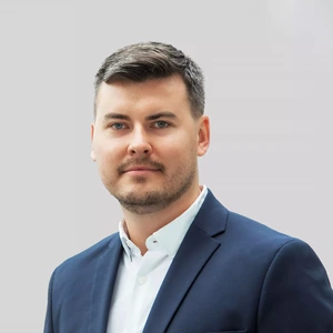 Portrait photo of a man with short hair and a light beard, wearing a white shirt and a dark blue blazer. He is looking directly at the camera with a slight smile. The background is light grey.