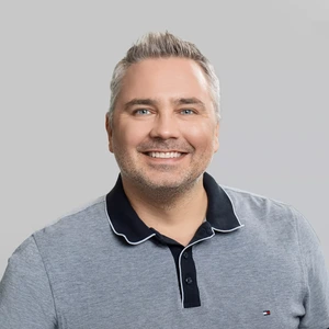 A man with greying hair and a short beard is wearing a grey polo shirt with a dark collar. He is smiling and looking at the camera against a light background.