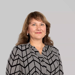 A woman with wavy brown hair is wearing patterned black and white zigzag shirt, pearl earrings, and is gently smiling while looking at the camera against a light background.