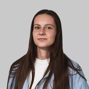 Portrait photo of a young woman with long dark hair, wearing a white T-shirt and light blue and white striped shirt. She is looking at the camera with a calm expression. The background is neutral light grey.