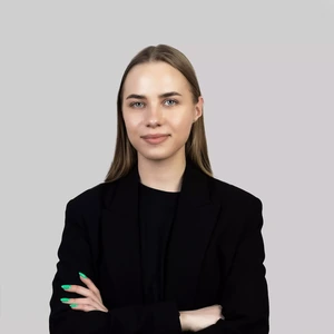 Portrait photo of a young woman with straight light hair, wearing a black blazer and top. She is standing with arms crossed, looking directly at the camera with a gentle smile. The background is light grey and neutral.