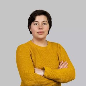 Portrait photo of a woman with short dark hair, wearing a mustard yellow sweater. She is standing with arms crossed and looking at the camera with a calm, composed expression. The background is neutral light grey.