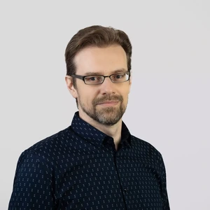 Portrait photo of a middle-aged man with short brown hair, a beard, and glasses. He is wearing dark patterned shirt and looks at the camera with a slight smile. The background is light gray and neutral.
