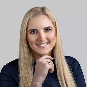 A woman with long blonde hair, wearing a dark shirt, smiles and gently rests her chin on her hand while looking at the camera against a light background.