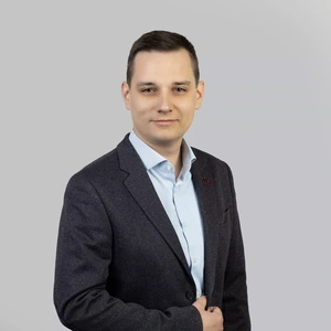 Portrait photo of a young man with short dark hair, wearing a light blue shirt and a dark blazer with a lapel pin. He is looking at the camera with a slight smile, against a light neutral background.