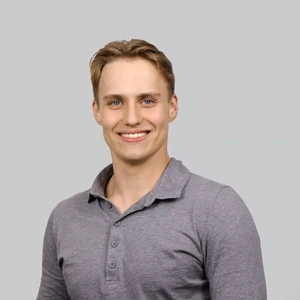 A young man with light hair, wearing a grey polo shirt, looking at the camera and smiling.