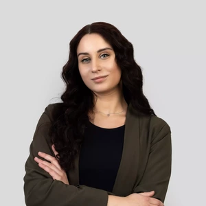 Portrait photo of a young woman with long dark wavy hair, wearing a black top and a dark green blazer. She is standing with arms crossed and looking at the camera with a confident expression. The background is neutral light grey.