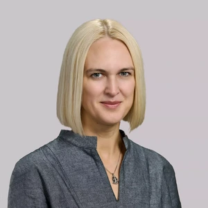 A blonde woman with straight hair, wearing a grey blouse with a stand-up collar, looking at the camera and smiling.