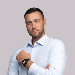 A serious-looking man with short dark hair, wearing a light blue shirt and a black wristwatch, posing with crossed arms against a light background.