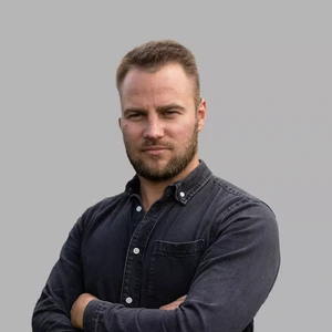 Portrait photo of a man with short light hair and a beard, wearing a dark denim-style button-up shirt. His arms are crossed, and he has a confident, serious facial expression. The background is grey.