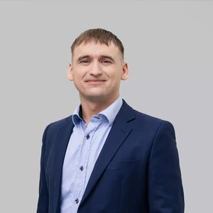 Portrait photo of a young man with short haircut, wearing a dark blue suit and a light blue shirt, smiling at the camera against a light background.